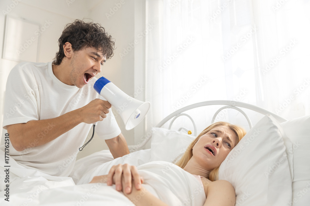 Obraz premium man using megaphone to wake up sleeping women in bed, funny and noisy morning scene. Concept of annoying wake-up call, couple humor, loud sound, relationship routine and domestic life.