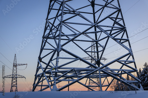 Snow-Covered High Voltage Power Pylons at Sunset in Winter Landscape, Industrial Infrastructure Against Cold Blue Sky and Orange Horizon, Energy Grid in Nature
