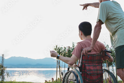 A young man with disability in wheelchair and his parent on the lake at sunset with background of the hill, Health care for the sick or person with disabilities, rehabilitation with natural therapy.