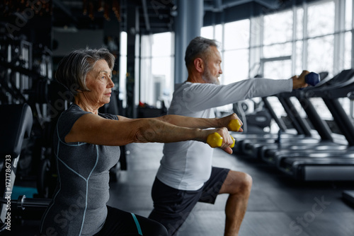 Elderly man and woman are doing squats with dumbbells in a gym