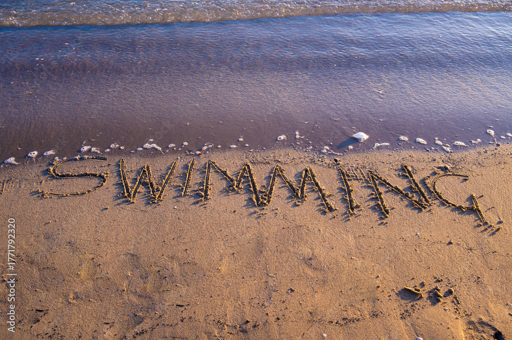 Fototapeta premium The word “Swimming” written on sandy beach near the shoreline