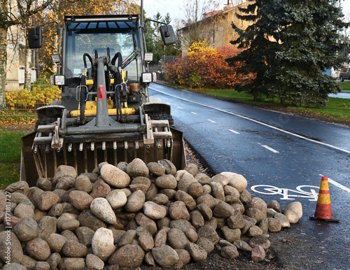 A front loader stands on a wet city road beside a bicycle lane, with a large pile of stones ready for construction or maintenance work