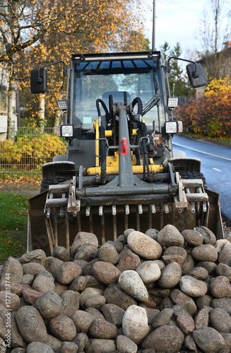 A front loader stands on a wet city road beside a bicycle lane, with a large pile of stones ready for construction or maintenance work