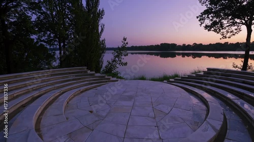 Amphitheater stone steps curve beside tranquil lake at dusk, purple sky reflecting on calm water, peaceful outdoor nature scene surrounded by trees