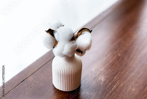 A white ceramic vase holds cotton branches with fluffy white bolls. The vase is placed on a wooden surface, creating a minimalist decor scene.