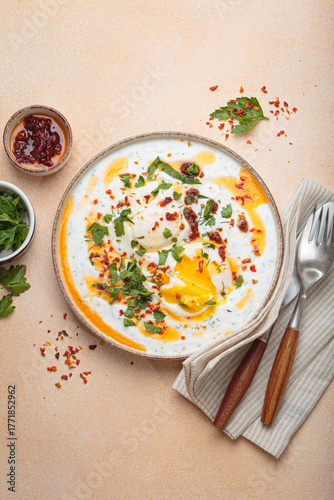 Turkish eggs cilbir, a delicious and colorful breakfast with poached eggs on yogurt, seasoned with spices and fresh herbs, arranged on a beige table