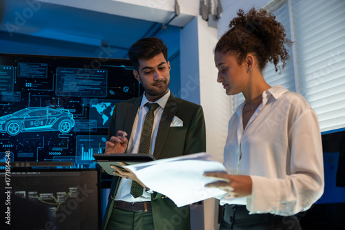 Two professionals analyzing and discussing documents with digital car interface in high-tech automotive innovation at dark modern office.