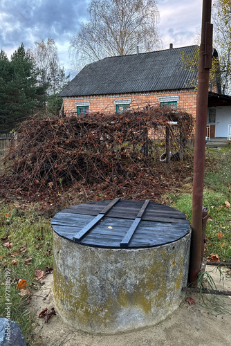 An unkempt vineyard. The vine is not pruned, part of it lies on the ground. In the foreground, a mine shaft; in the background, a residential house.