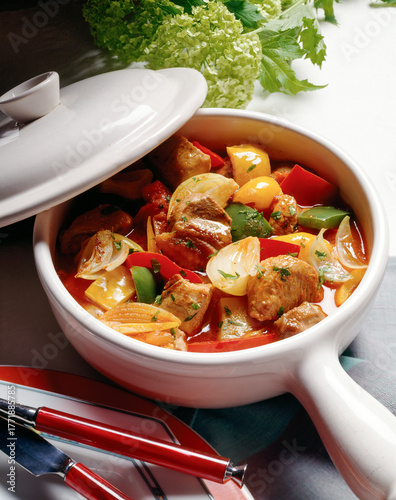 A Delicious stew featuring chicken meat, vibrant vegetables and herbs served in a white dish. The meal is ready to serve on a table with fresh greens nearby