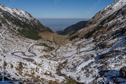 panoramic view of the Transfagarasan Highway winding through the snowy peaks of the Carpathian Mountains in Romania. Scenic mountain road under bright sunlight, famous travel destination in Europe.