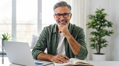 Caucasian man with grey hair and beard wearing glasses and green shirt smiling, posing at camera while using laptop and notebook in modern home office. Remote work and happy lifestyle with copy space
