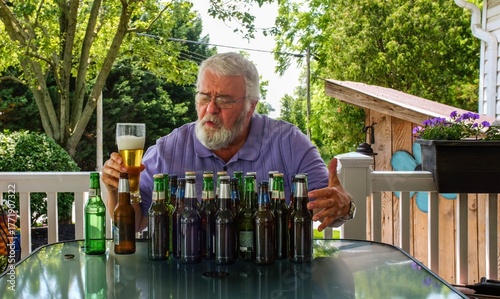 Fotografija An elderly man holds a glass of beer and examines several empty beer bottles on a table
