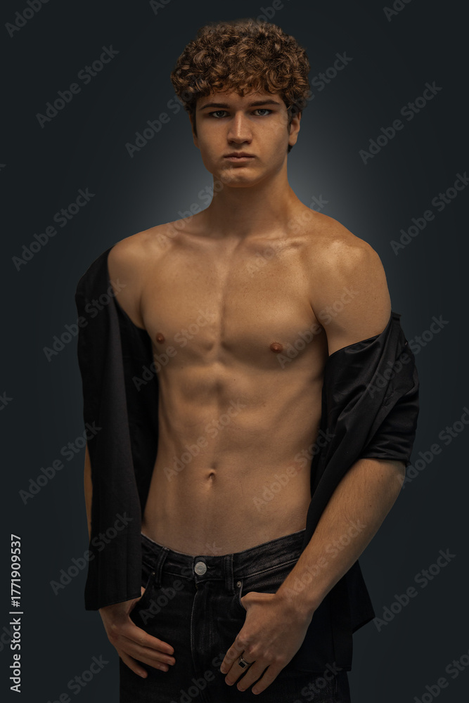Fototapeta premium young man stands in a studio showcasing his curly hair and toned physique. He wears a black shirt casually draped over one shoulder while looking directly at the camera.