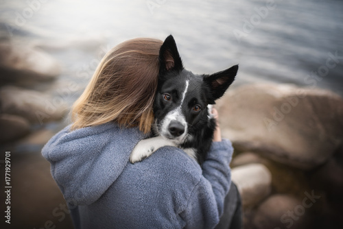 A woman with a border collie dog enjoys a peaceful moment by the sea. The black and white dog leans into her embrace as gentle waves lap at the shore, creating a serene atmosphere