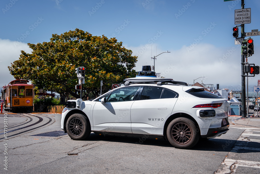 Obraz premium A white Waymo SUV with lidar and sensors waits at a light by cable car tracks in San Francisco, near Hyde Street and Beach Street, in cool midday foggy light.