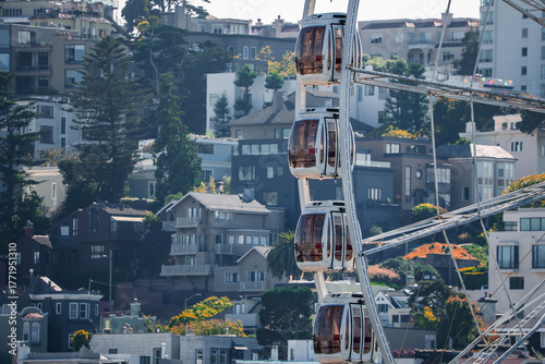 Close up of glass gondolas ascending beside San Francisco hillside homes and steep streets in bright midday light, white wheel contrasts textured urban forms.