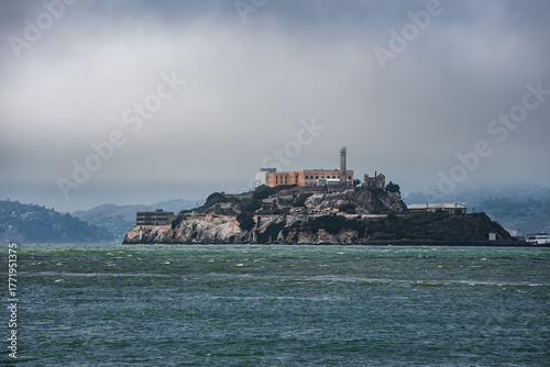 Rocky Alcatraz Island rises from choppy waters in San Francisco Bay, with the main prison and lighthouse visible under overcast light, wide horizontal view.