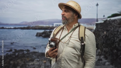 Man holding vintage camera and gesturing with hand on seaside street wearing pith helmet and backpack; adventure curiosity.