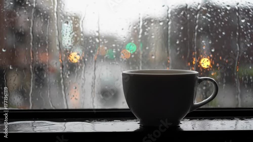 A white mug sits on a wet surface with rain streaming down a window, soft city lights in background