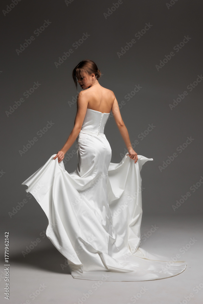 Naklejka premium Elegant bride poses in minimalist white satin wedding gown against soft natural light backdrop