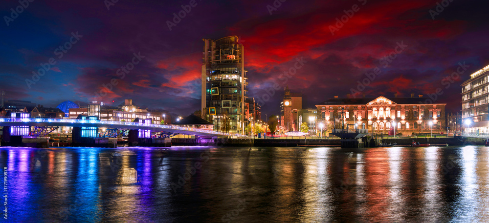 Fototapeta premium Belfast City night skyline with the Lagan Weir Pedestrian Bridge and colorful reflections on the River Lagan in Northern Ireland, United Kingdom, tranquil and vibrant green space riverwalk landscape.