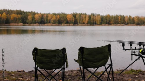 Two empty fishing chairs by calm autumn lake with rods on tripod, peaceful outdoor recreation scene with forest reflection and tranquil atmosphere, leisure and relaxation in nature