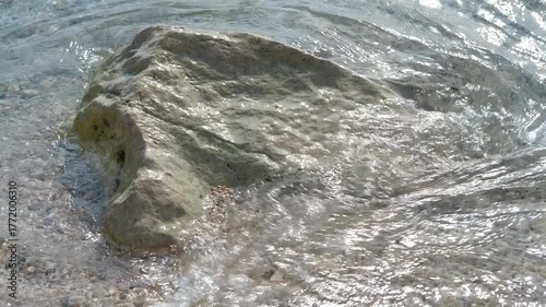 Gentle waves washing over coastal rock on sandy beach. Crystal-clear water gently washing over large, textured rock resting on sandy shore, revealing serene coastal landscape with soft waves