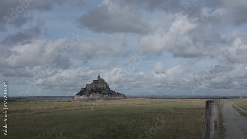 Time lapse video of tidal fields to Mont Saint-Michel, the iconic island abbey in Normandy, France. The stone monastery rises dramatically under a wide, cloud-filled sky.