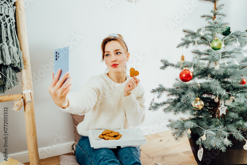 Happy Young Woman in cosy sweater taking selfie photo while relaxing on floor cusions near potted christmas tree in modern Scandi interior home. Cozy winter holidays. Content creating. Selective focus