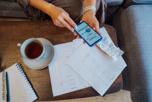 Managing personal finances on smartphone. No face woman checking financial balance on smartphone while enjoying a cup of tea at home. Financial documents, receipts on the table. focused on budgeting.