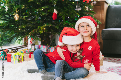 Two smiling kids in festive sweaters and Santa hats sitting under Christmas tree at home during holiday. Happy sister cuddling brother, feeling excited, having fun, childhood miracle time concept