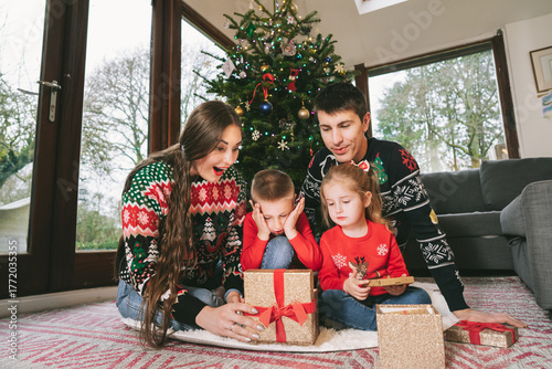 Happy family in festive jumpers sitting under Christmas tree at decorated home. Emotional parents and kids feeling excited of unwrapping Christmas gifts, having fun, Family time, childhood concept