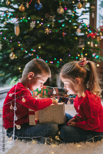 Two smiling kids in festive sweaters sitting under Christmas tree at home. Emotional sister and brother feeling excited of unwrapping Christmas gifts, having fun, childhood miracle time concept.