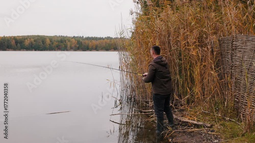 Fisherman casting spinning rod while predator fishing, man standing near reeds on autumn lake, outdoor sport fishing action and nature recreation concept with calm water and forest reflection