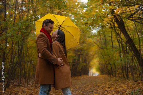 Romantic couple standing under yellow umbrella in autumn park, smiling and embracing among colorful fall trees, wearing matching brown coats and enjoying cozy outdoor moment.
