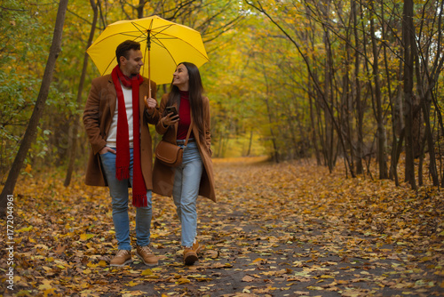 Smiling young couple walking under a yellow umbrella in an autumn park, dressed in brown coats and red accents, enjoying a romantic stroll surrounded by colorful fall leaves.