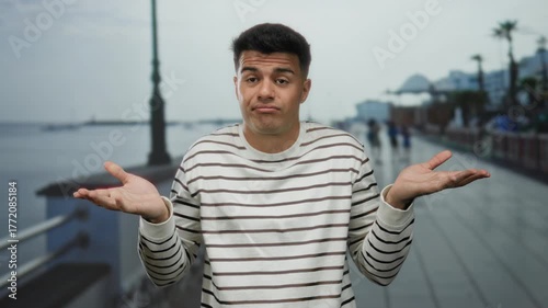 Young man gestures with expression on a seaside promenade showing a relaxed and humorous demeanor captured against a serene beach background.