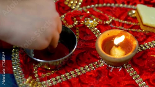 A hand performing a Hindu ritual with a lit diya and red powder on a decorative red cloth.