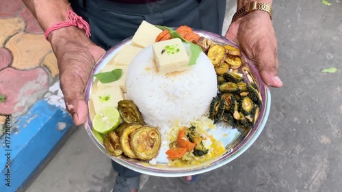 A person holds a plate of traditional Indian vegetarian food, featuring a mound of rice topped with butter and surrounded by various cooked vegetables.
