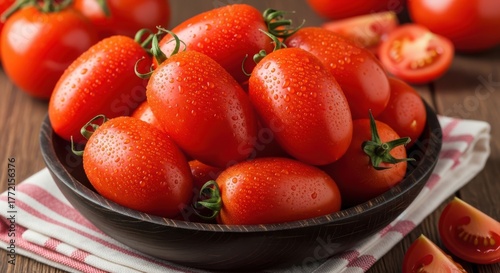 A dark bowl filled with ripe, red roma tomatoes, some with water droplets, on a wooden surface