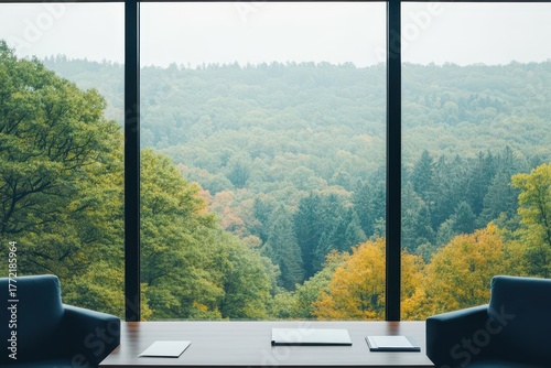 Modern office interior with large panoramic window overlooking lush green forest and rolling hills, minimalistic workspace with two chairs and a sleek table