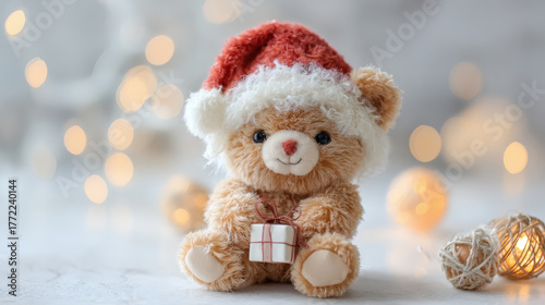 Cute teddy bear wearing a Santa hat holding a small gift box with festive bokeh lights in the background.