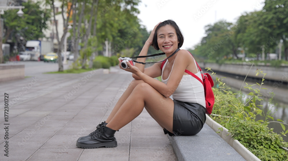 Fototapeta premium A cheerful Asian woman with short hair wearing a leather mini skirt and combat boots sits outdoors by a canal, smiling and holding her small digital camera