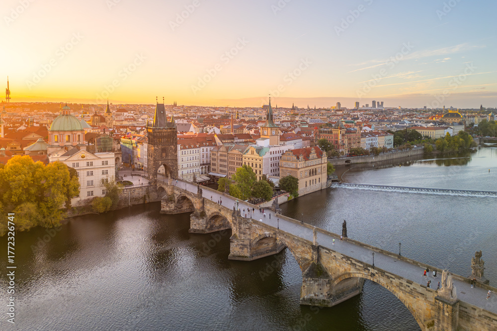 Naklejka premium Morning sunlight gently illuminates Prague Castle in the background, while people stroll across Charles Bridge over the Vltava River, creating a serene early day scene in Prague.