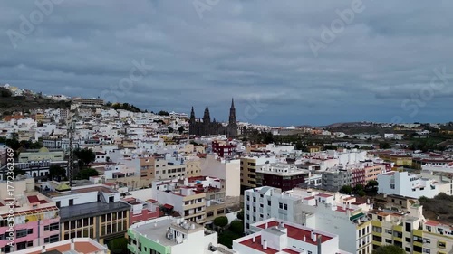 aerial view of the town of Arucas, Gran Canaria, featuring the prominent, dark Gothic Revival Church of San Juan Bautista rising above the dense cluster of white and colored traditional buildings.