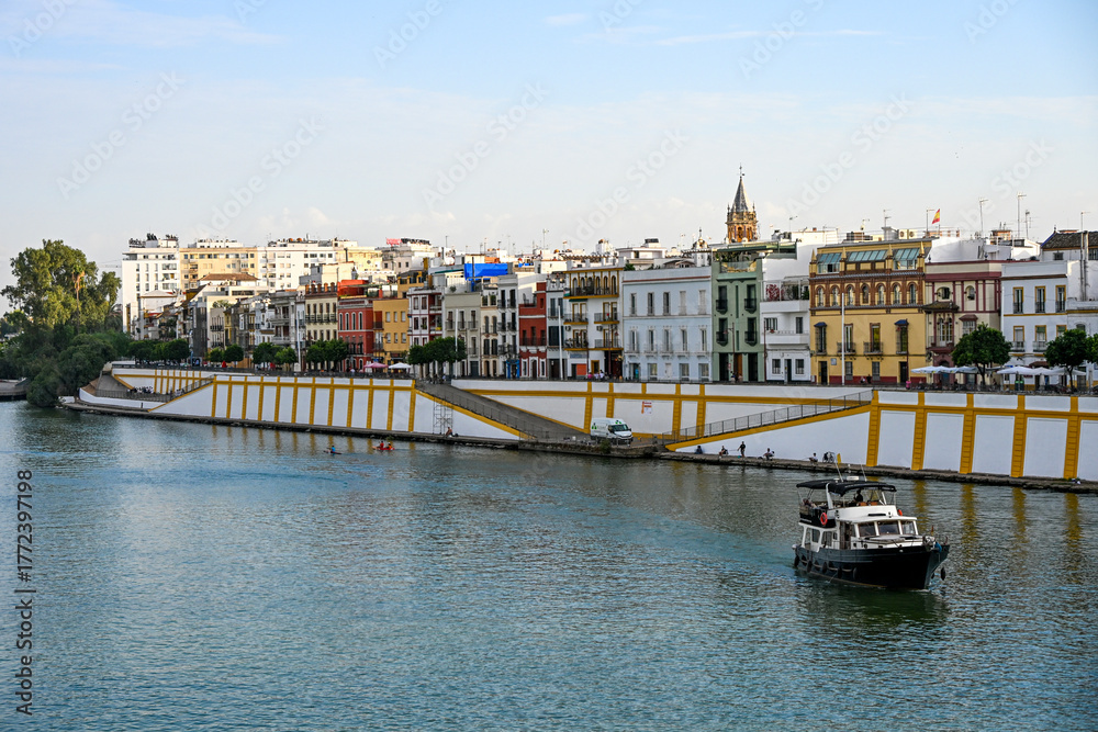 Obraz premium Colourful riverside buildings of the Triana district reflected on the Guadalquivir River in Seville, with a boat gliding calmly through the water