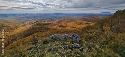 Wide panorama from Tar-kő peak in the Bükk Mountains, Hungary. Rolling forested hills covered in golden and orange autumn foliage under dramatic clouds.
