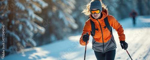 Young woman in orange jacket cross country skiing on snowy trail. Female athlete during winter sport activity in forest. Sportswoman with poles practices ski on sunny day.