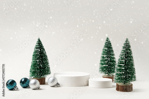 Winter holiday product mockup featuring miniature fir trees, white podiums and assorted ornaments with gentle snowfall - stylish seasonal display for product and gift visuals.