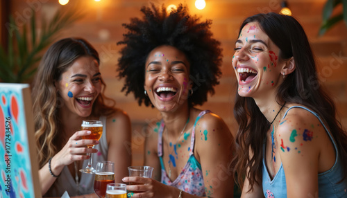 Fototapeta Naklejka Na Ścianę i Meble -  Three diverse women laugh loudly at paint, sip event. They have colorful paint on faces, clothes. Friends enjoy art therapy, drinks, creative time together. Happy girls socialize, create art.
